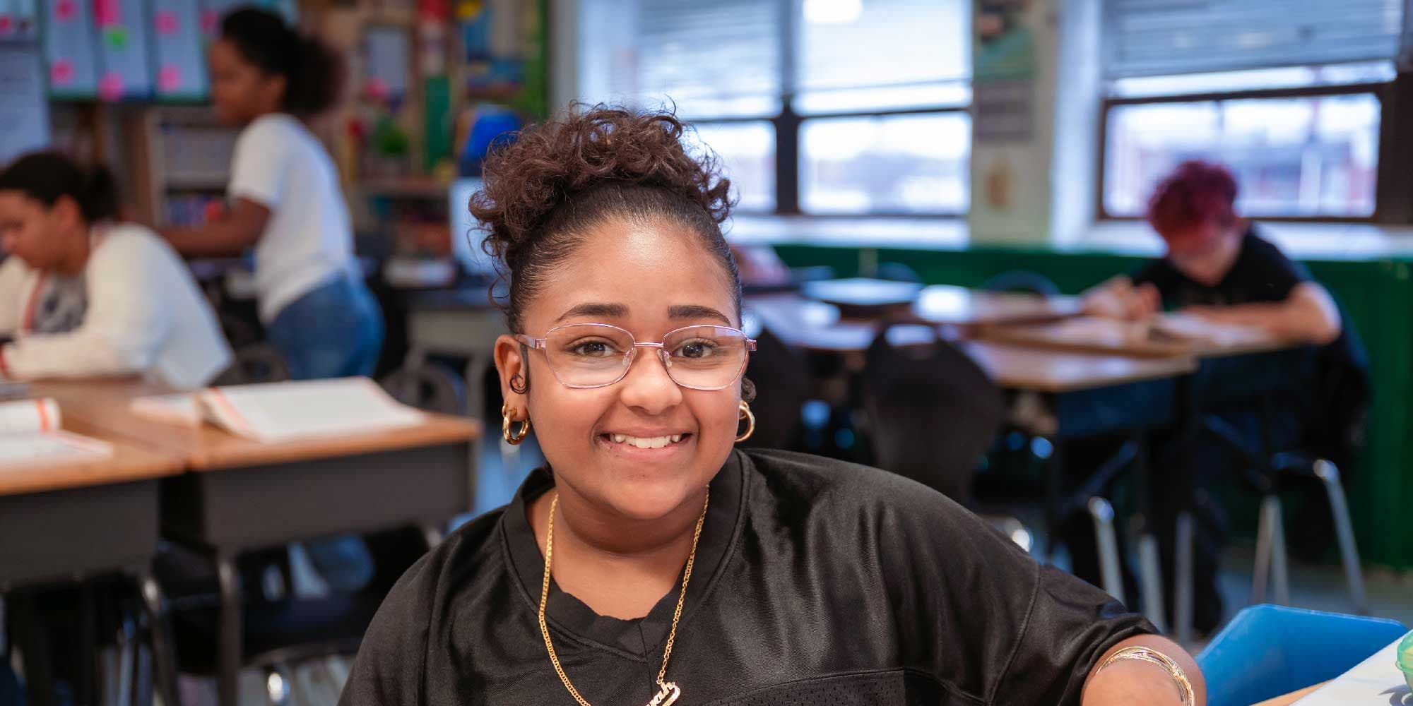 Smiling student in glasses at a classroom desk.