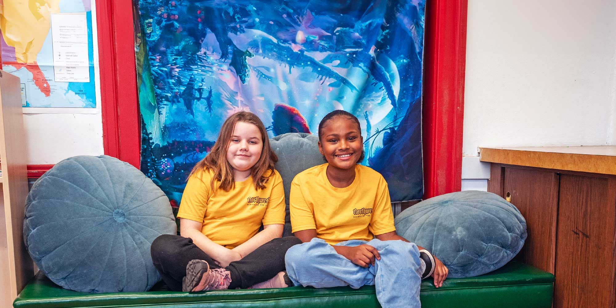 Two smiling students in yellow Northwest School of the Arts T-shirts sit on a classroom bench.